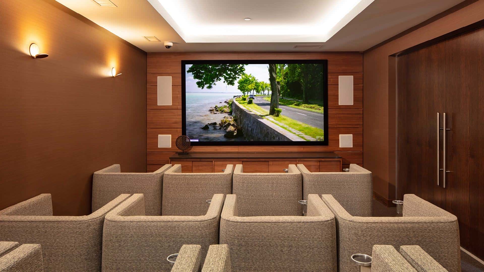 Home theater with tiered beige seating rows, wood-paneled walls, recessed lighting, and wall-mounted flat-screen TV displaying a lakeside scene.