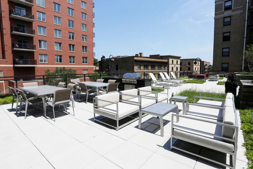 Rooftop terrace with white patio furniture, dining tables, lounge seating, and grill, surrounded by brick apartment buildings.