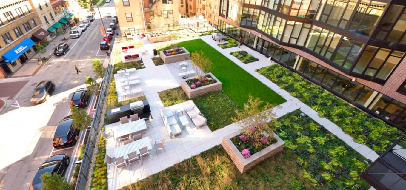 Aerial view of rooftop terrace with green lawn, raised planter beds, white lounge furniture, and surrounding Evanston street and buildings.