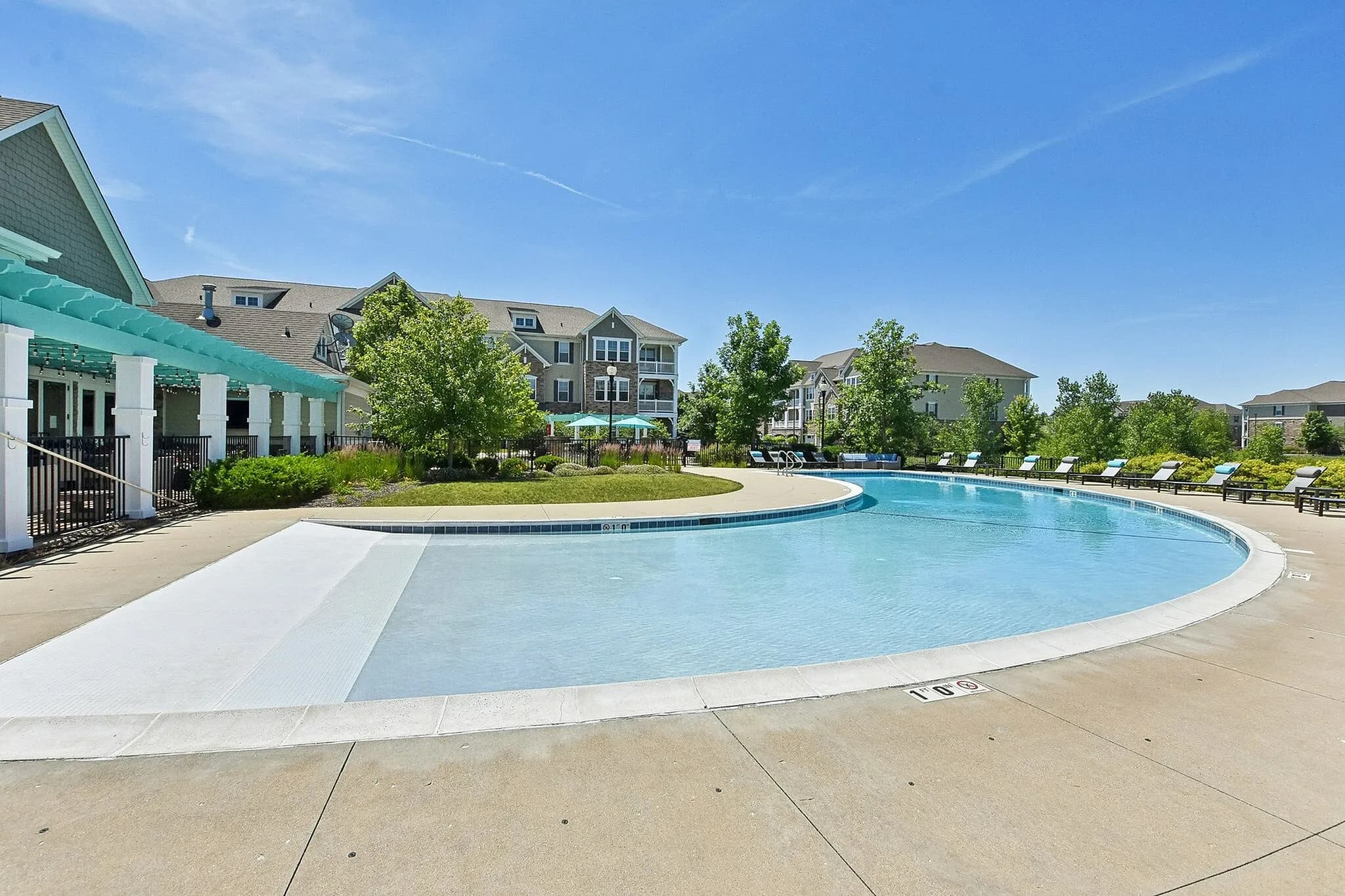 Curved resort-style swimming pool with clear blue water, concrete deck, white pergola structure, and multi-story residential buildings in background under clear sky.