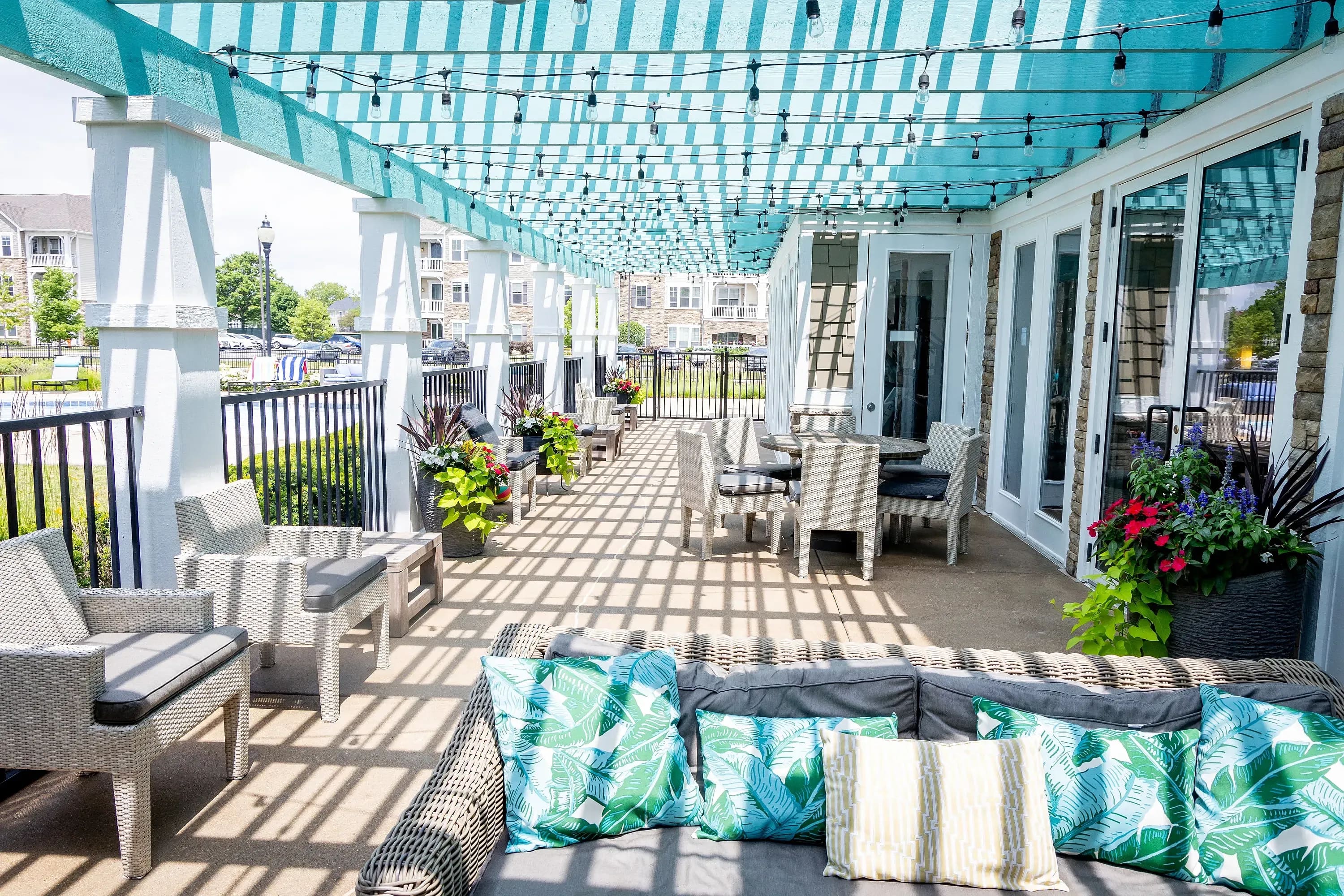 Covered outdoor patio with turquoise striped pergola, white wicker seating, tropical patterned cushions, and waterfront buildings visible beyond railing.