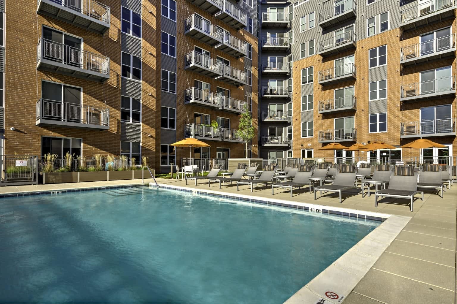 Outdoor swimming pool with gray lounge chairs and yellow umbrellas, flanked by multi-story brick apartment buildings with balconies.