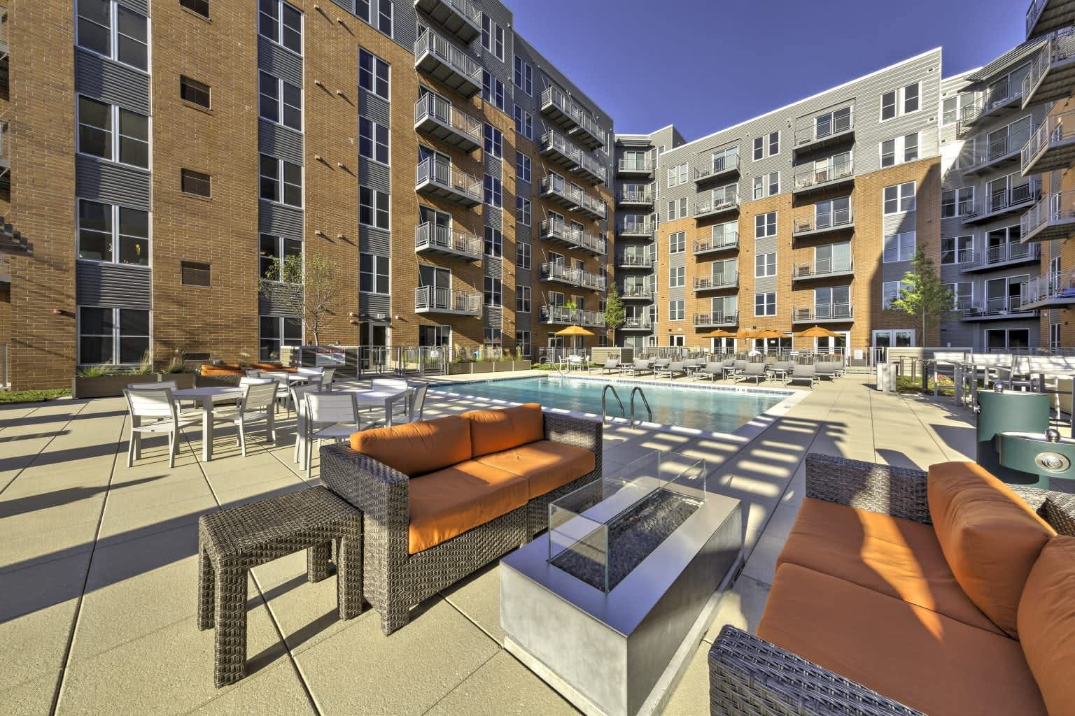 Outdoor pool courtyard with woven lounge chairs, fire pit, and surrounding brick apartment buildings under clear sky.