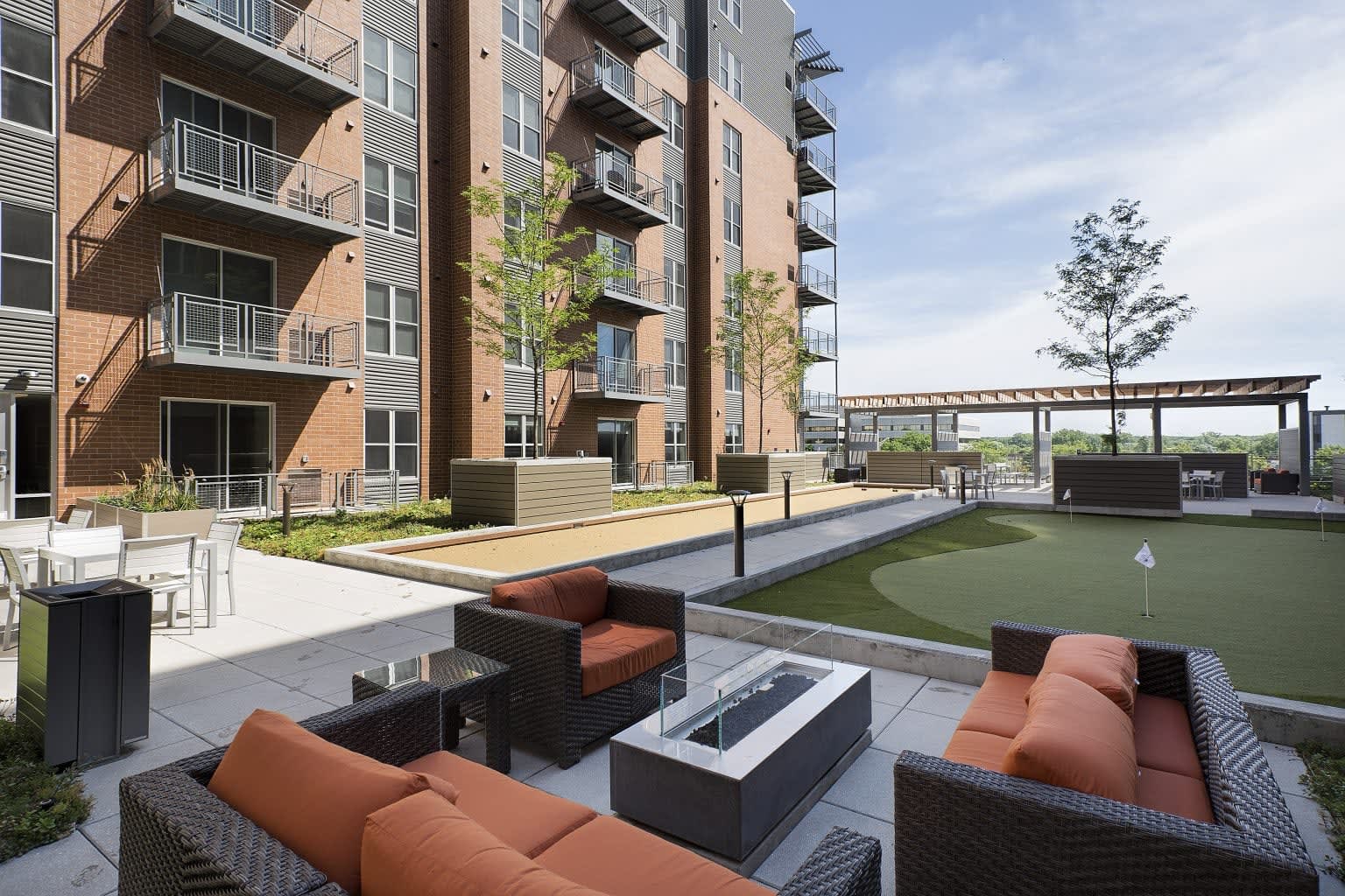 Rooftop terrace with wicker lounge seating, fire pit, bocce court, putting green, and multi-story brick building backdrop.