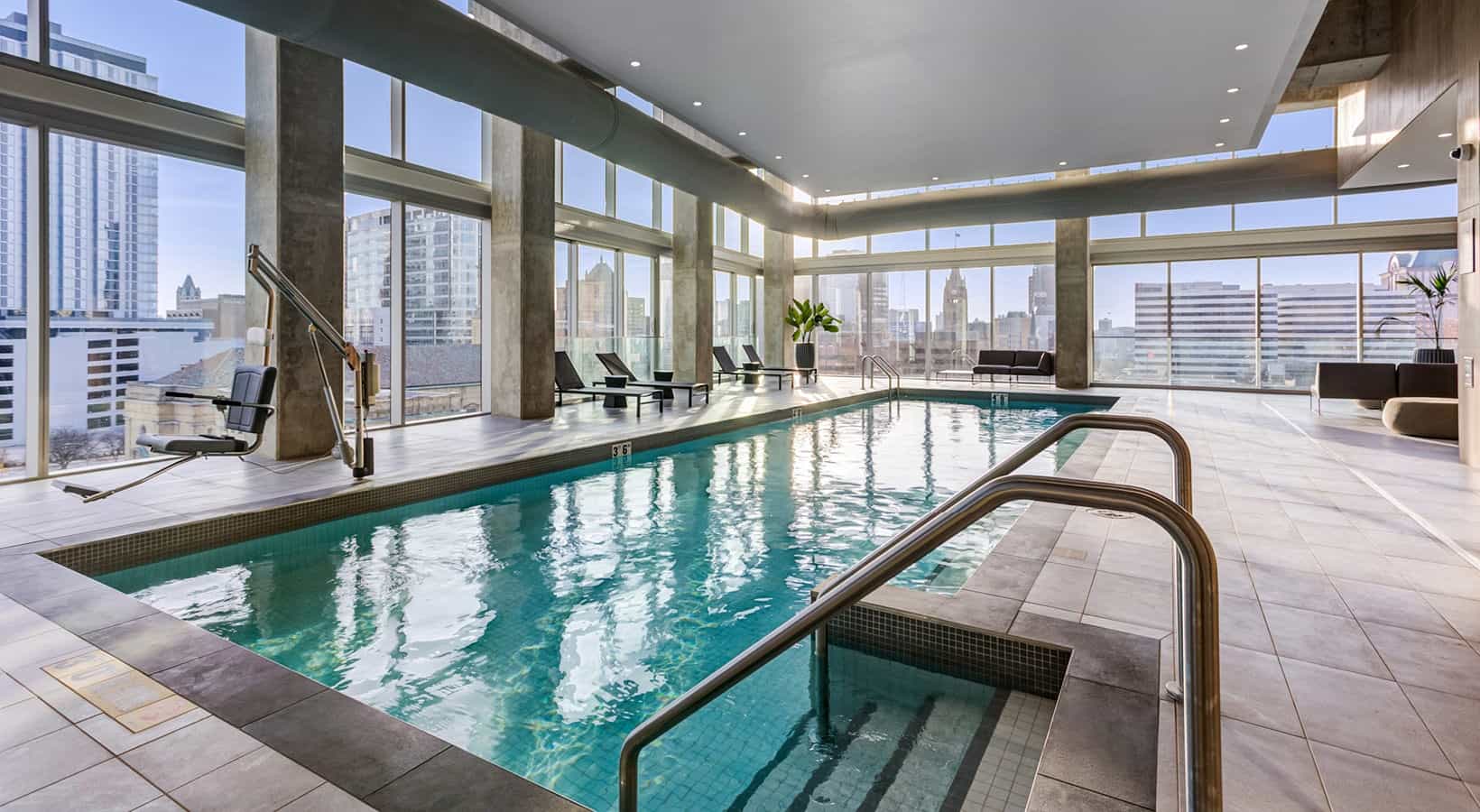 Indoor swimming pool with metal handrails, surrounded by lounge chairs and floor-to-ceiling windows with views of downtown Milwaukee buildings.