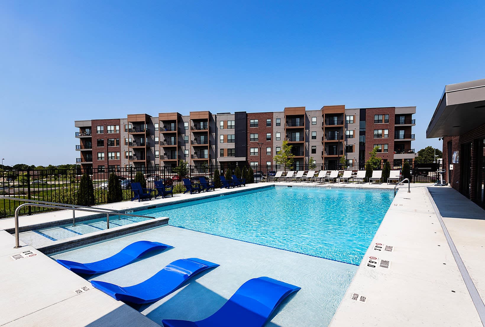 Resort-style swimming pool with blue lounge chairs, shallow sun shelf, and multi-story brick apartment building in background under clear sky.