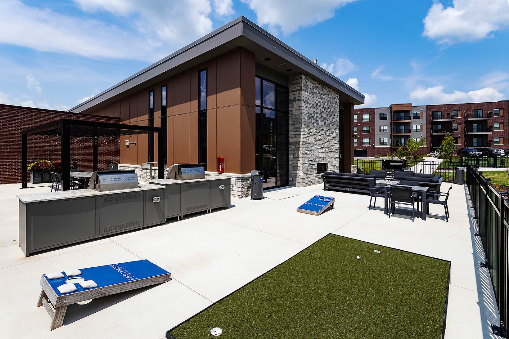 Modern rooftop terrace with outdoor kitchen, dining tables, cornhole boards, putting green, and contemporary building with stone and brown metal cladding.