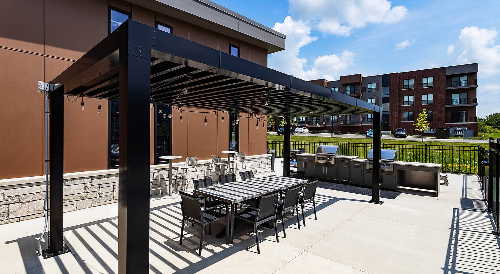 Rooftop patio with black pergola, dining table, lounge seating, built-in grill, and brown shade screens overlooking green lawn and brick apartment buildings.
