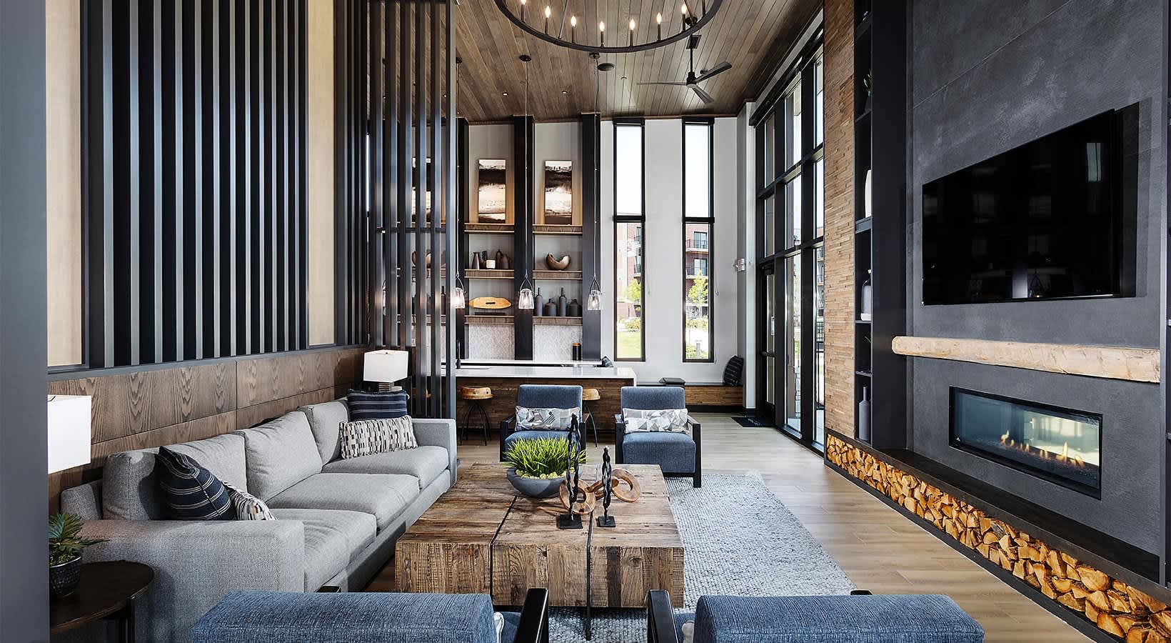 Modern living room with gray sectional sofa, wood ceiling, fireplace, wall-mounted TV, and large windows with black frames.