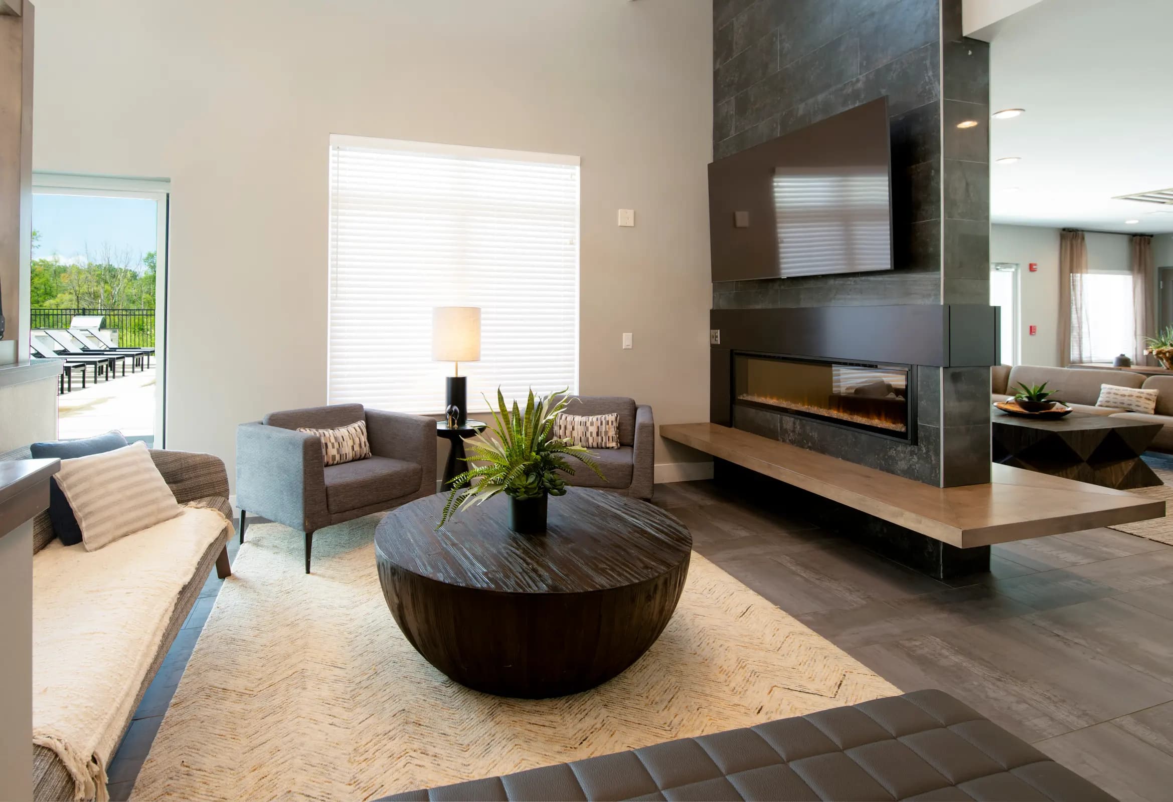 Modern living room with brown leather chairs, round black coffee table, wall-mounted TV above linear fireplace, and large window with white blinds.