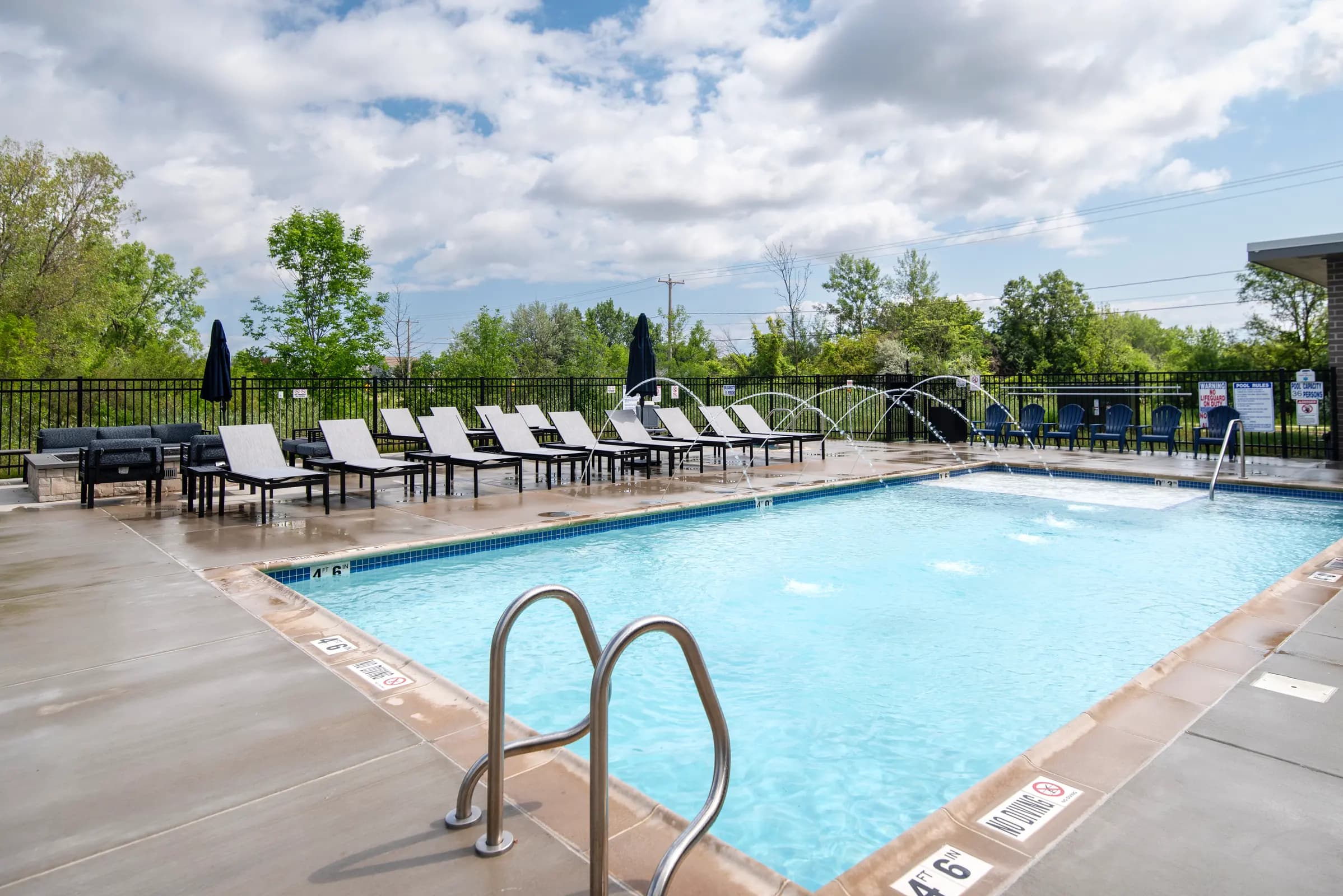 Resort pool with clear blue water, metal ladder in foreground, lounge chairs and umbrellas on surrounding deck, trees and fencing in background.