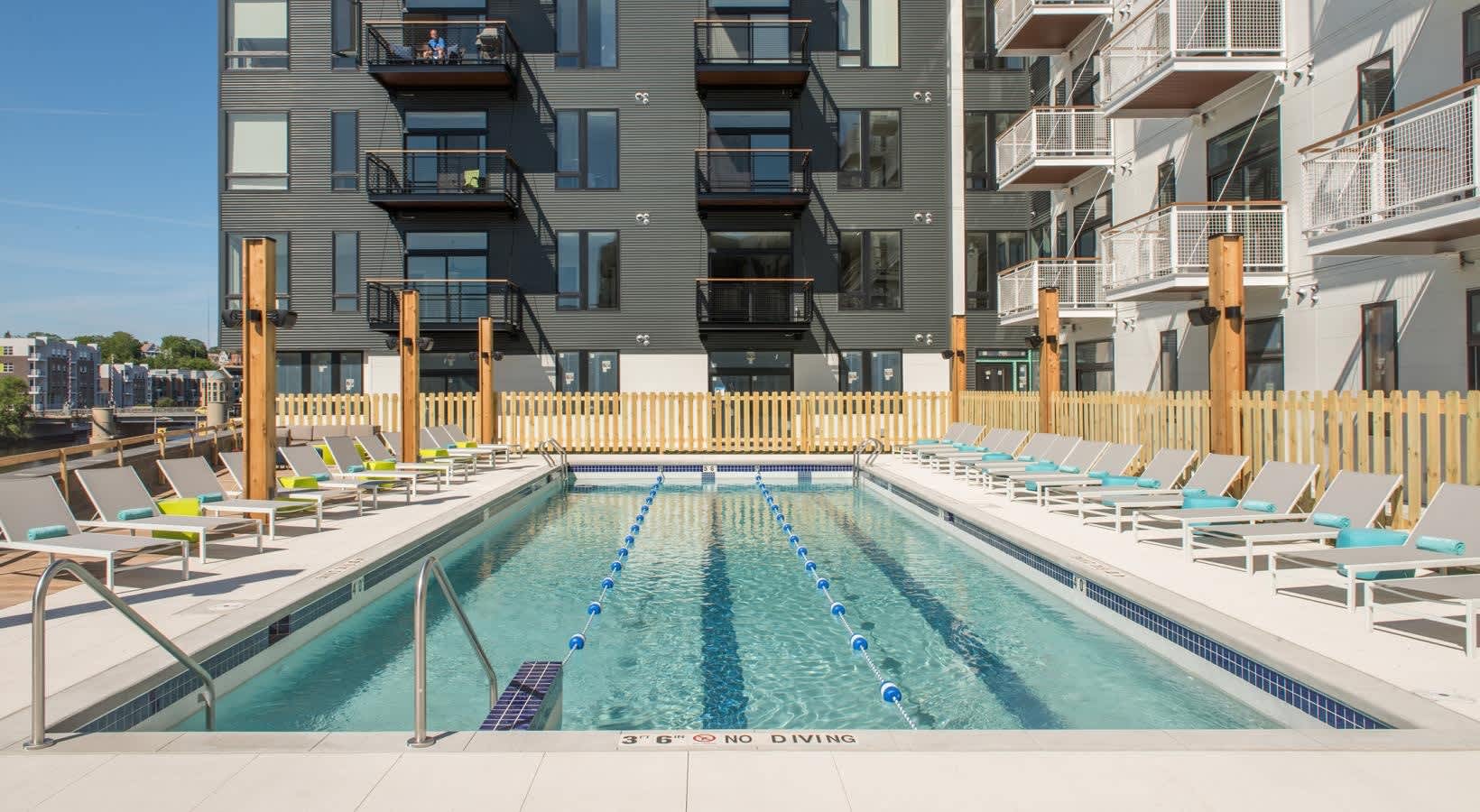 Lap pool with blue lane dividers, lounge chairs on both sides, wooden fence, and multi-story apartment buildings in background.