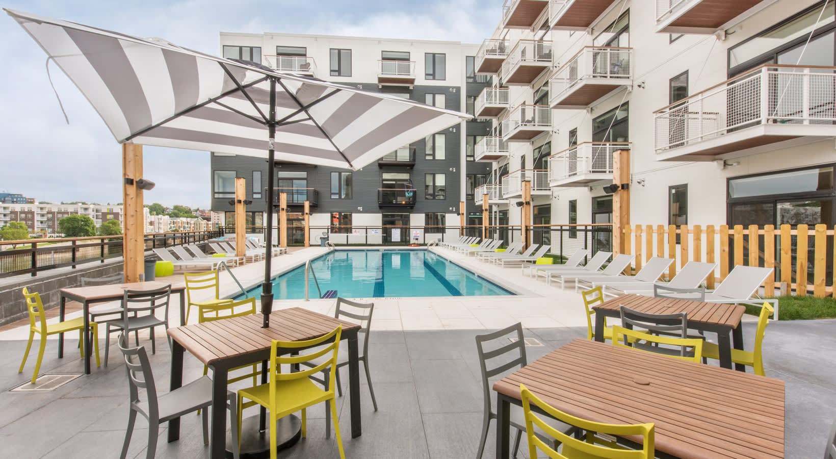 Resort-style pool deck with striped umbrella, wooden dining tables, yellow chairs, and multi-story residential buildings in background.