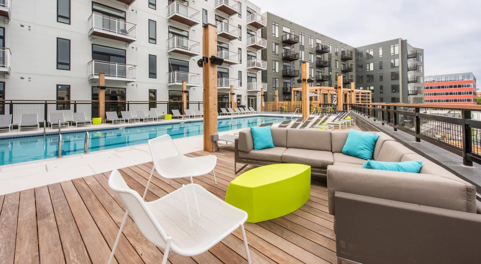 Rooftop pool deck with wooden decking, lounge chairs, gray sectional sofa with blue pillows, lime green ottoman, and resort-style pool surrounded by multi-story apartment buildings.