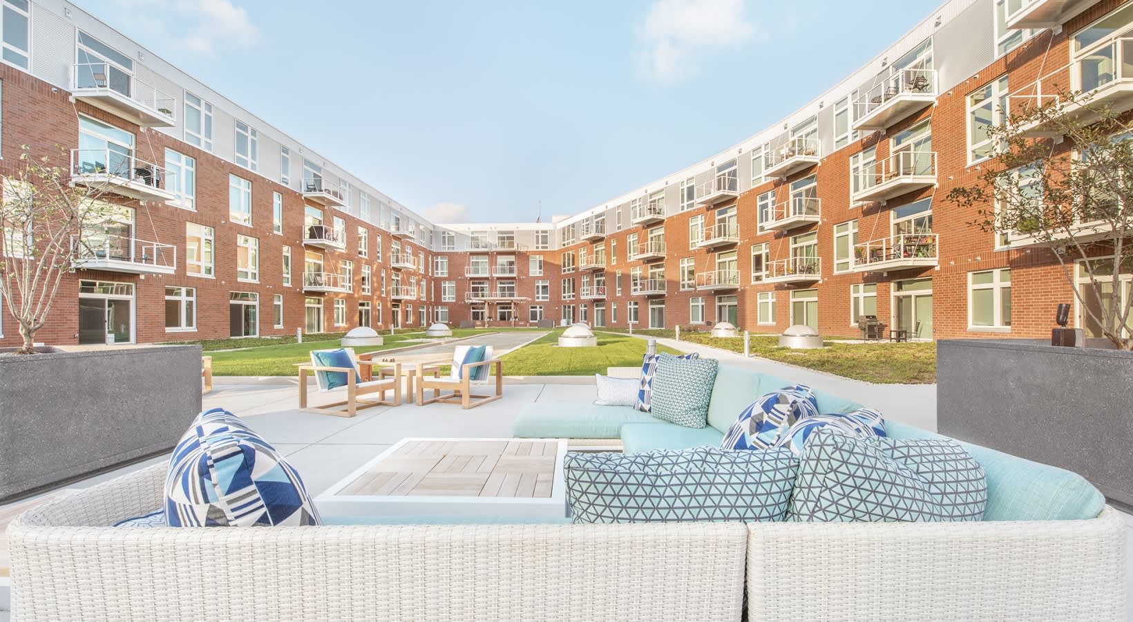 Woven outdoor seating with blue and white pillows overlooking a central courtyard with green lawn, wooden deck, and multi-story brick apartment buildings.