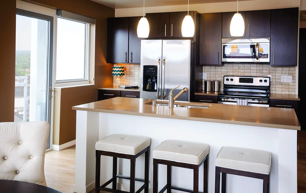 Contemporary kitchen with dark wood cabinets, stainless steel refrigerator and range, white subway tile backsplash, pendant lights, and white island with three upholstered stools; large windows show city views.