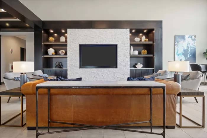 Living room with cognac leather sofa, wall-mounted TV, white brick accent wall, and dark shelving with decorative objects.