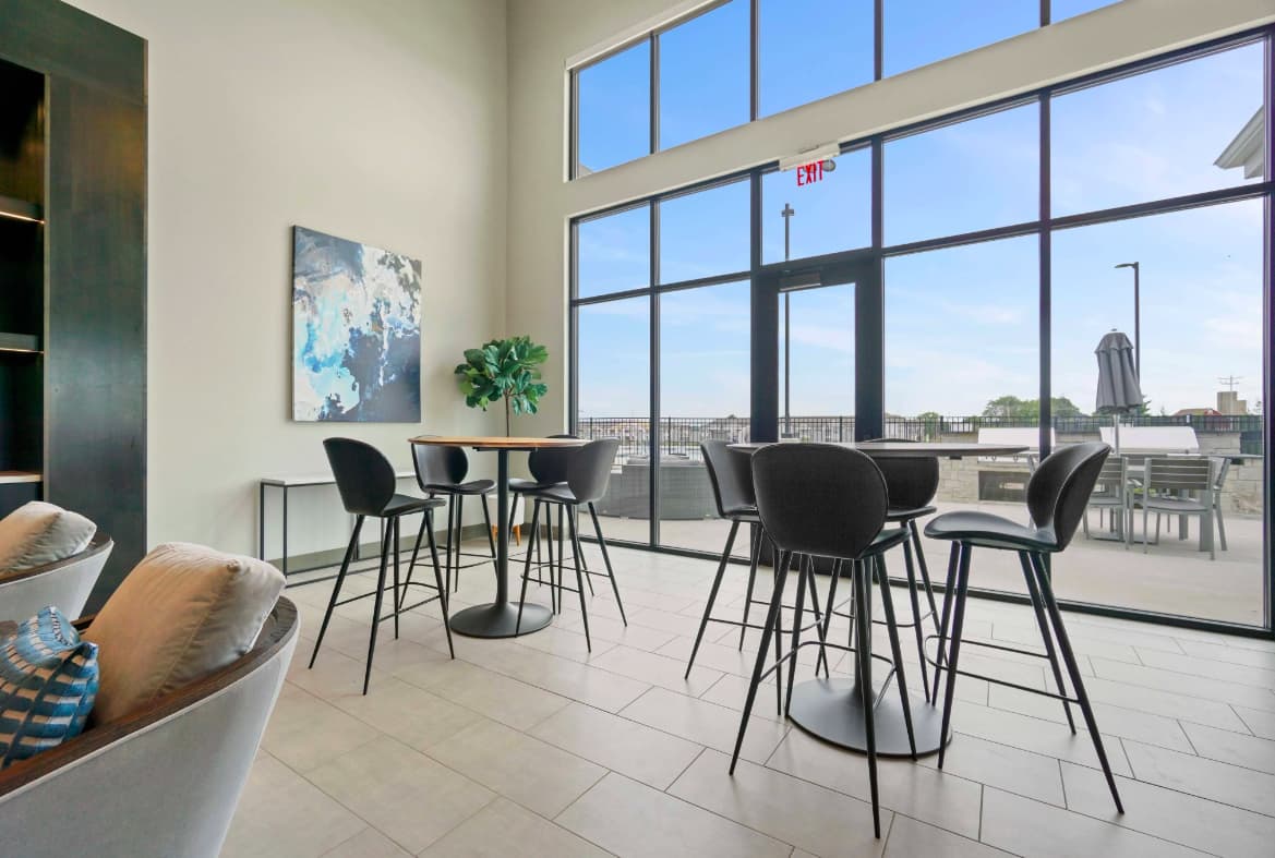 High-rise lounge with black bar stools, round table, floor-to-ceiling windows overlooking cityscape, light tile flooring, and contemporary furnishings.