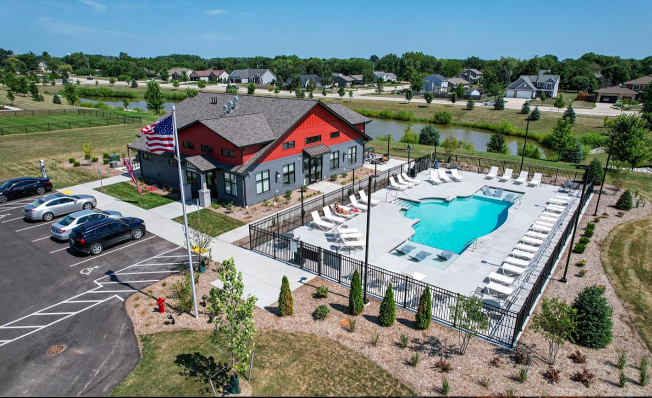 Aerial view of red and gray clubhouse with American flag, fenced resort pool with white lounge chairs, parking area, and surrounding residential community.