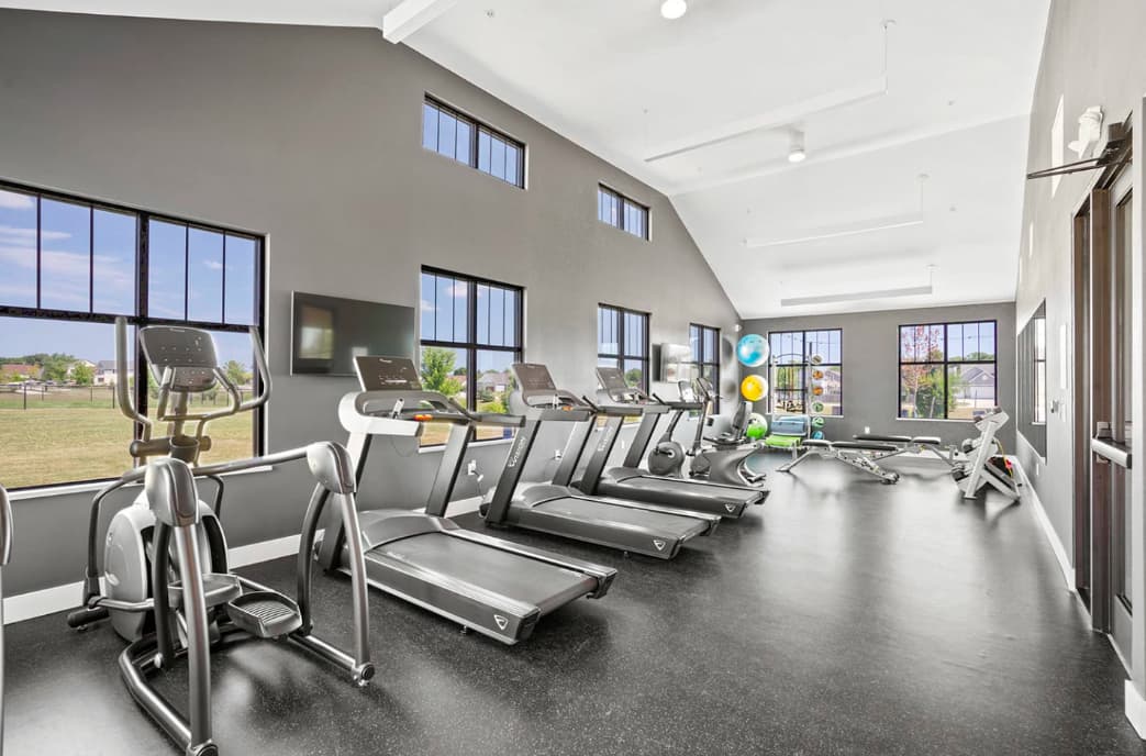 Modern fitness center with rows of treadmills and exercise bikes, large windows, white vaulted ceiling, and dark flooring.