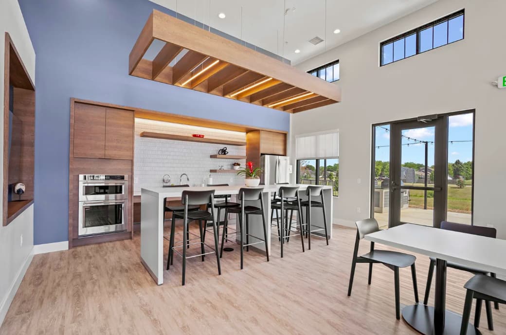 Modern kitchen with wood cabinetry, white island with bar seating, stainless steel appliances, and open dining area with large windows overlooking green landscape.