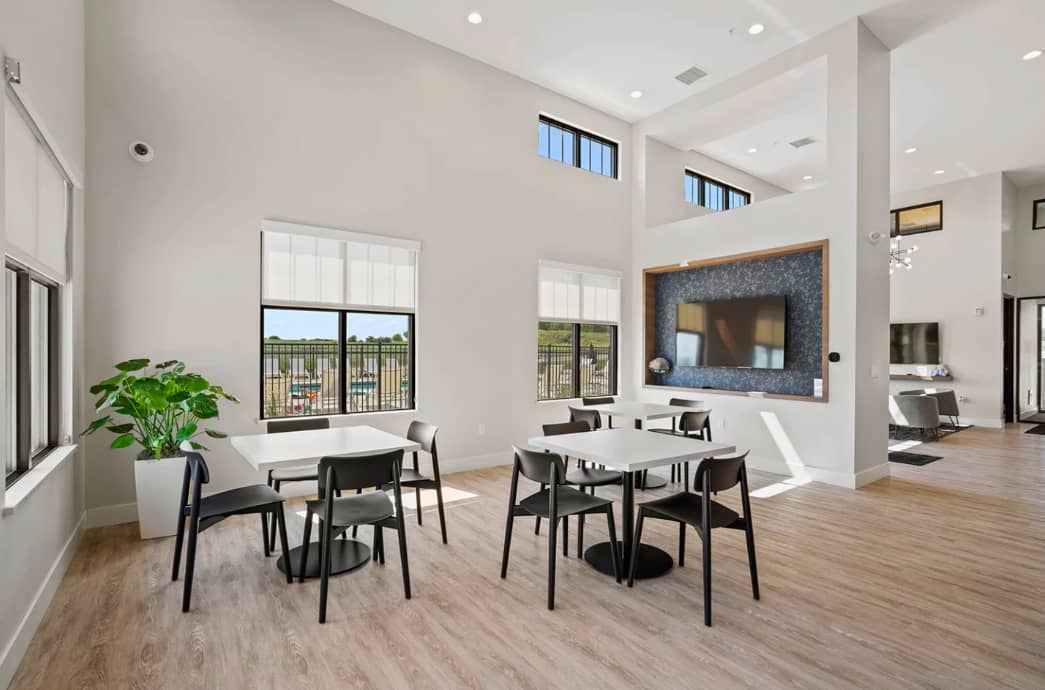 Open-concept dining area with two white tables, black chairs, potted plant, large windows with field views, and wall-mounted TV.