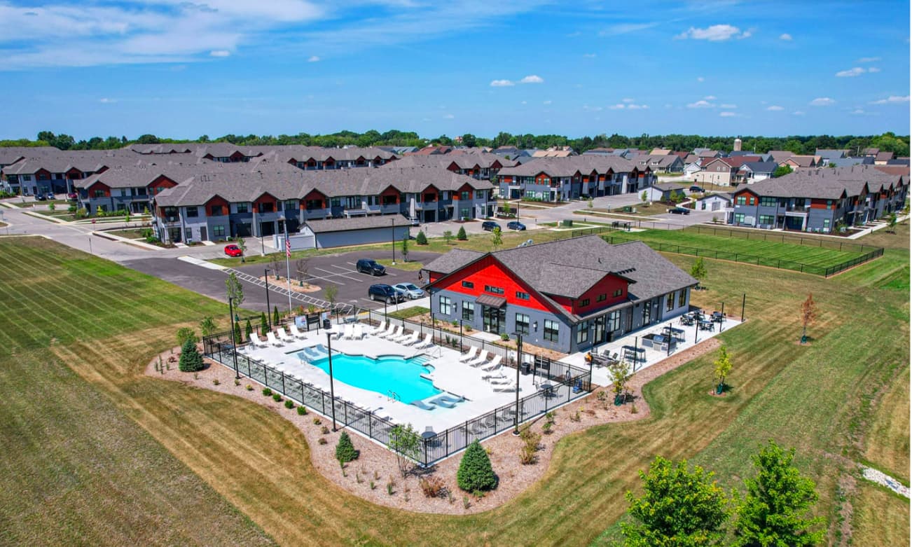 Aerial view of community clubhouse with red roof and turquoise swimming pool surrounded by lounge chairs, with residential buildings and green lawns in background.