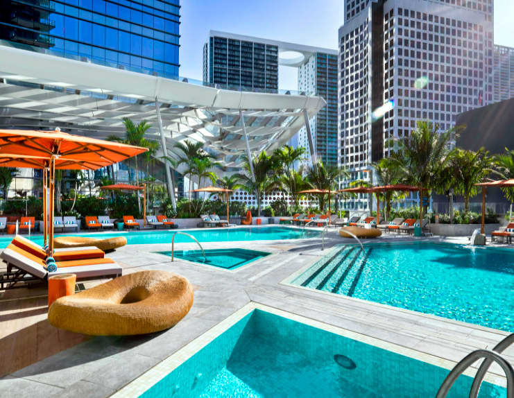 Rooftop pool deck with turquoise pools, orange umbrellas, lounge chairs, palm trees, and downtown Miami high-rises in background.