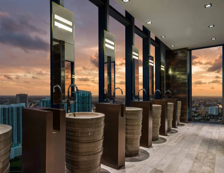 Modern bathroom with multiple vanities, tall mirrors, and floor-to-ceiling windows overlooking Miami skyline at sunset.