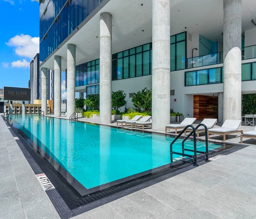 Turquoise lap pool with lounge chairs, white concrete columns, and modern building facade with green-tinted windows.