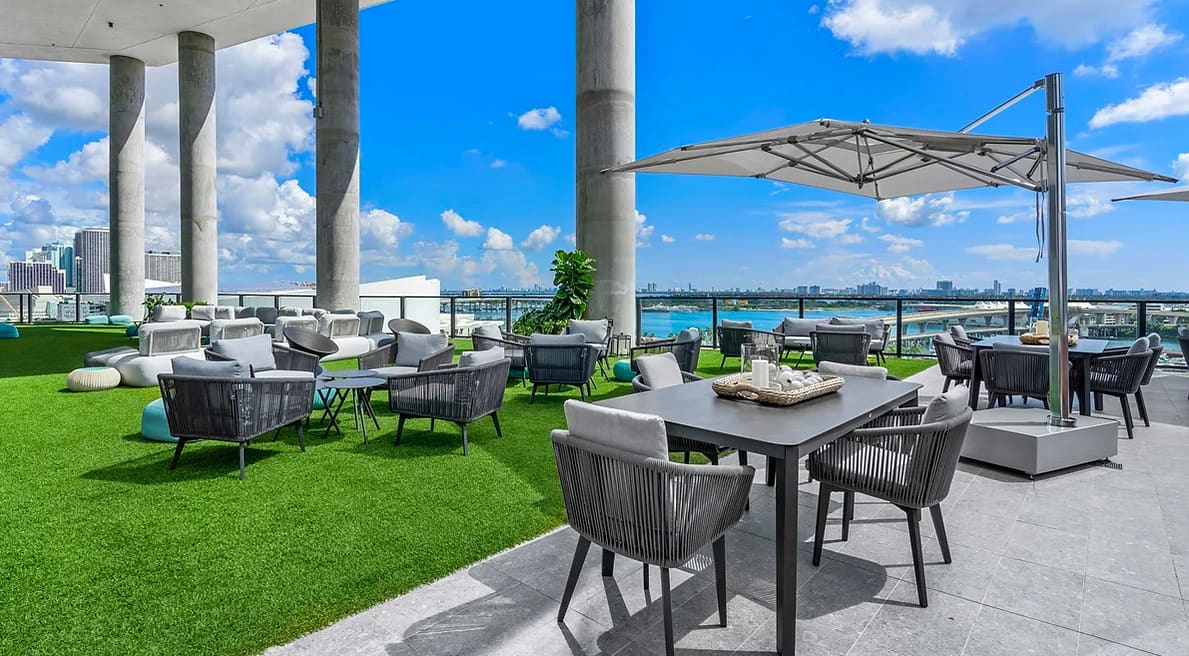 Rooftop terrace with dining table, lounge seating, cantilever umbrellas, artificial turf, and panoramic waterfront views of Miami and Biscayne Bay.