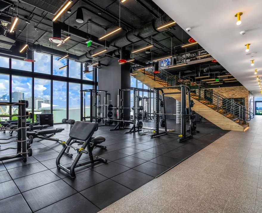 Fitness center with exercise machines, black rubber flooring, large windows overlooking Miami skyline, industrial ceiling with warm lighting, and wooden staircase.