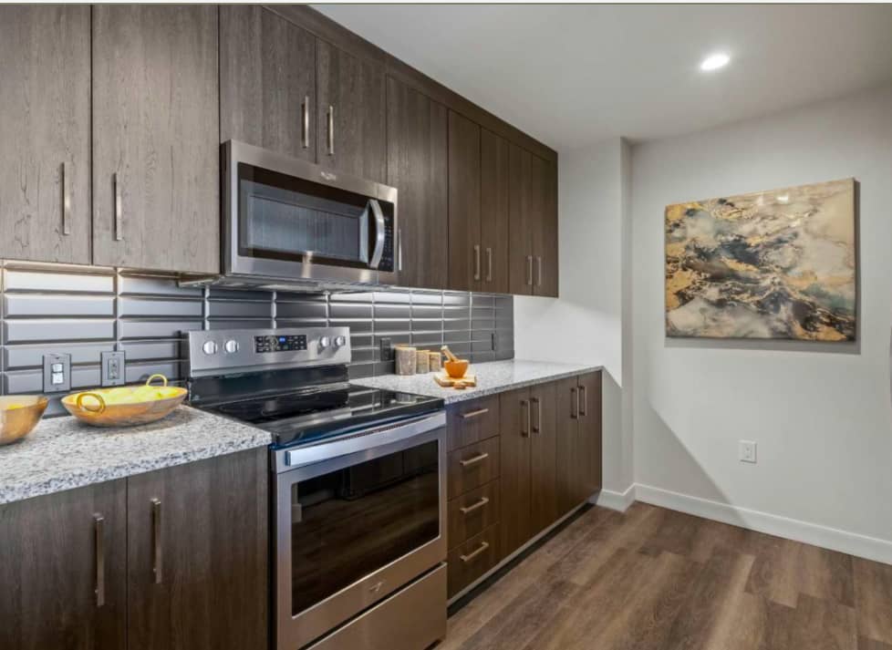 Kitchen with dark wood cabinets, stainless steel range and microwave, gray subway tile backsplash, granite countertop, and wood-look flooring.