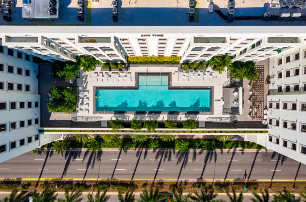 Overhead view of rectangular turquoise pool with white lounge chairs, palm trees, and surrounding multi-story apartment buildings in urban setting.