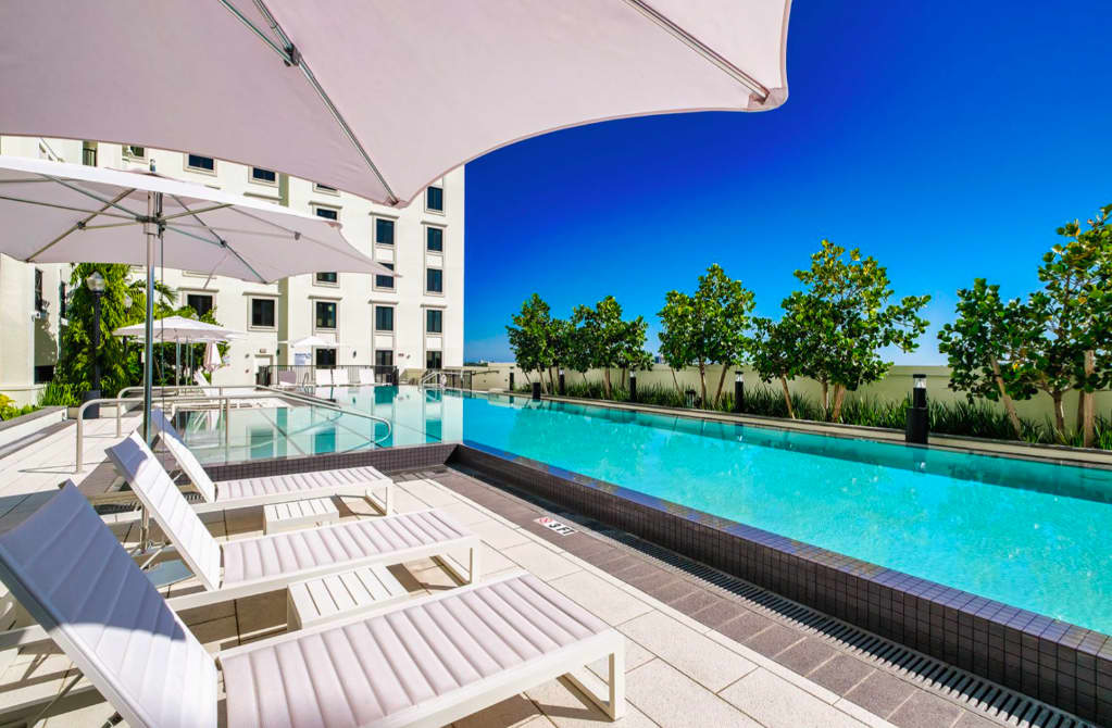 Resort pool with turquoise water, white loungers, umbrellas, and modern white apartment building in background under clear blue sky.