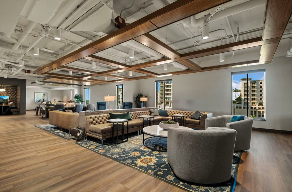 Open-concept lounge with tan leather sectional, gray armchairs, wood beam ceiling, and large windows with city views.
