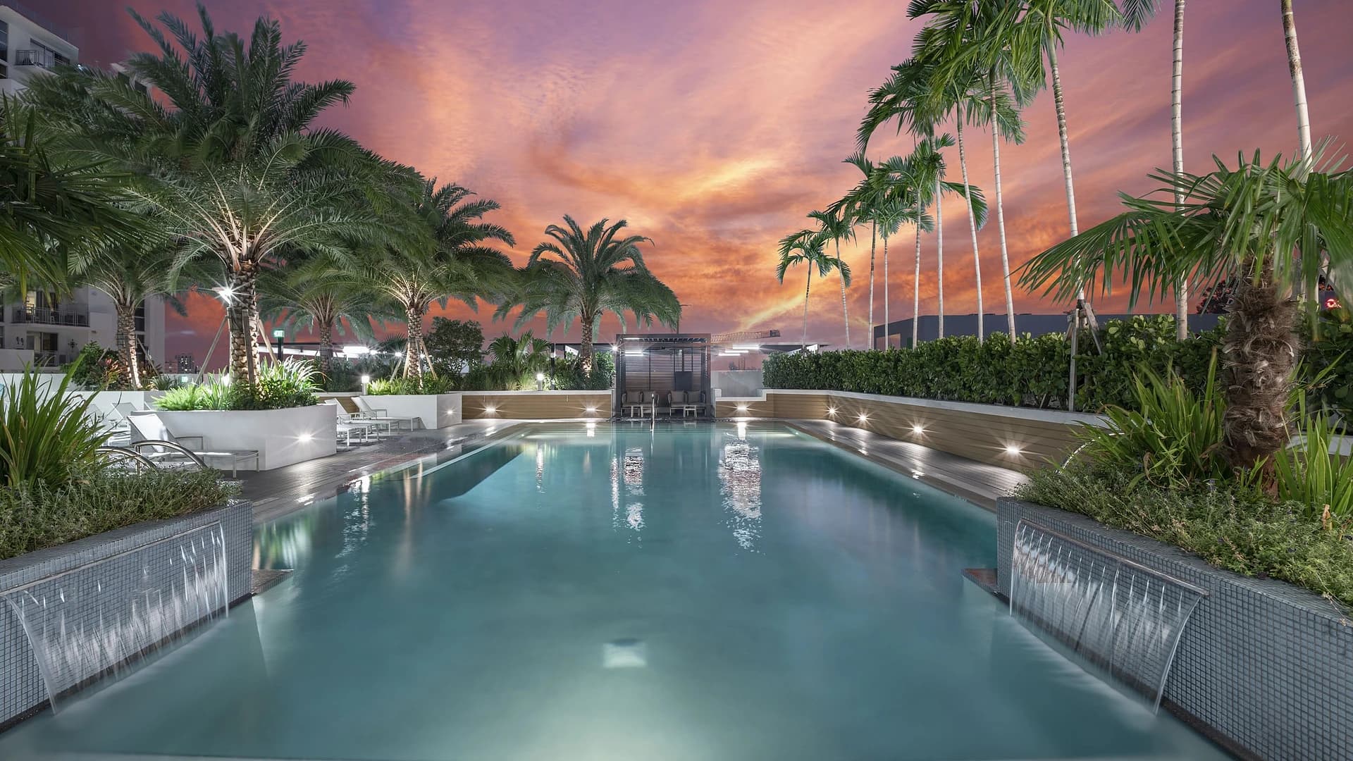 Illuminated resort pool surrounded by palm trees and white lounge chairs at dusk, with colorful sunset sky overhead.