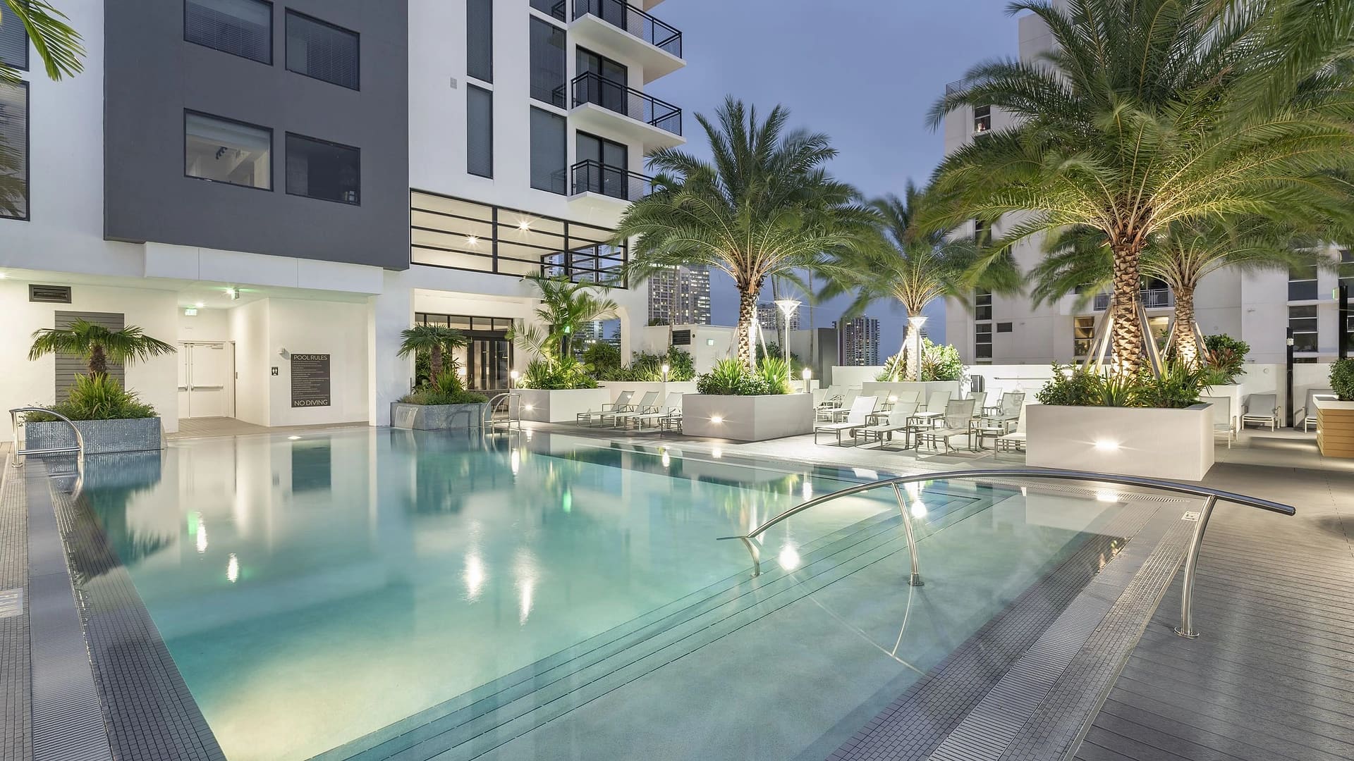 Resort-style pool courtyard with clear turquoise water, palm trees, white lounge chairs, and modern white residential buildings at dusk.