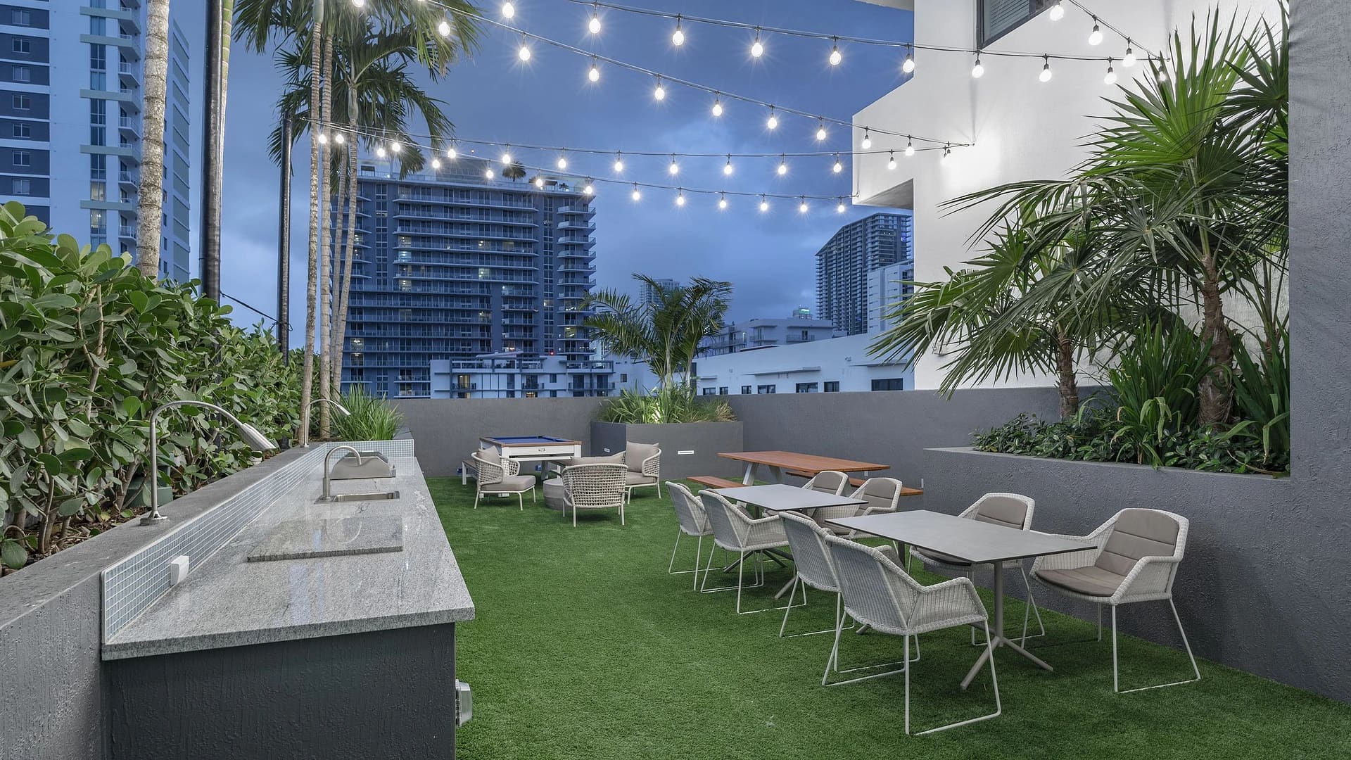 Rooftop terrace with artificial turf, dining table, lounge seating, potted palms, string lights, and downtown Miami skyline at dusk.