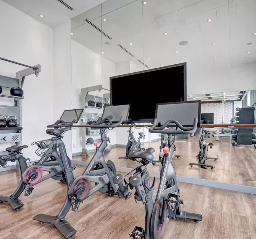 Modern fitness studio with multiple stationary bikes, wall-mounted screens, mirrored walls, and wood flooring.