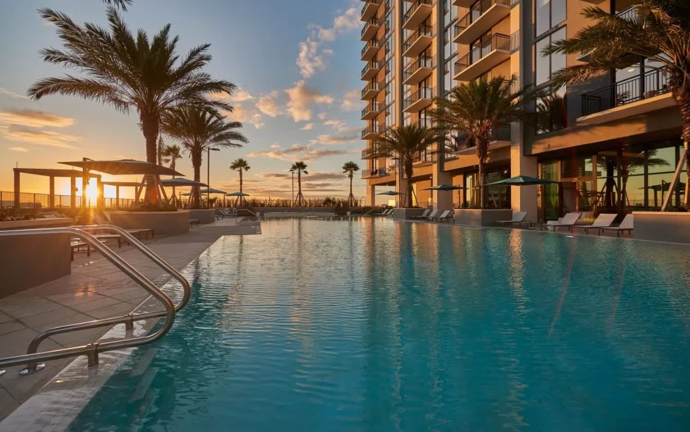 Resort pool at sunset with turquoise water, palm trees, lounge chairs, and multi-story residential building in Miami.