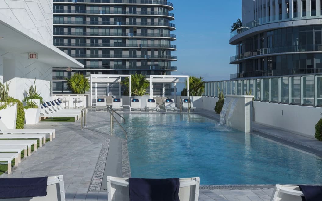 Rooftop pool with clear blue water, white lounge chairs, cabanas, and palm plants overlooking downtown Miami buildings under clear sky.