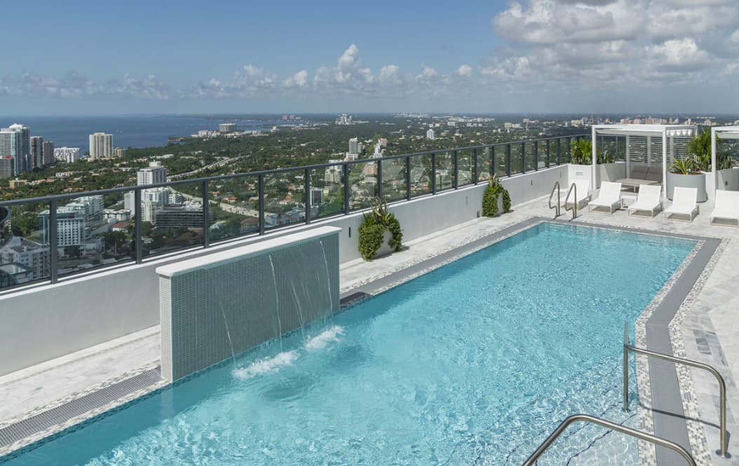 Rooftop pool with clear turquoise water, white lounge chairs, glass railing, and expansive views of Miami's coastline and downtown skyline.