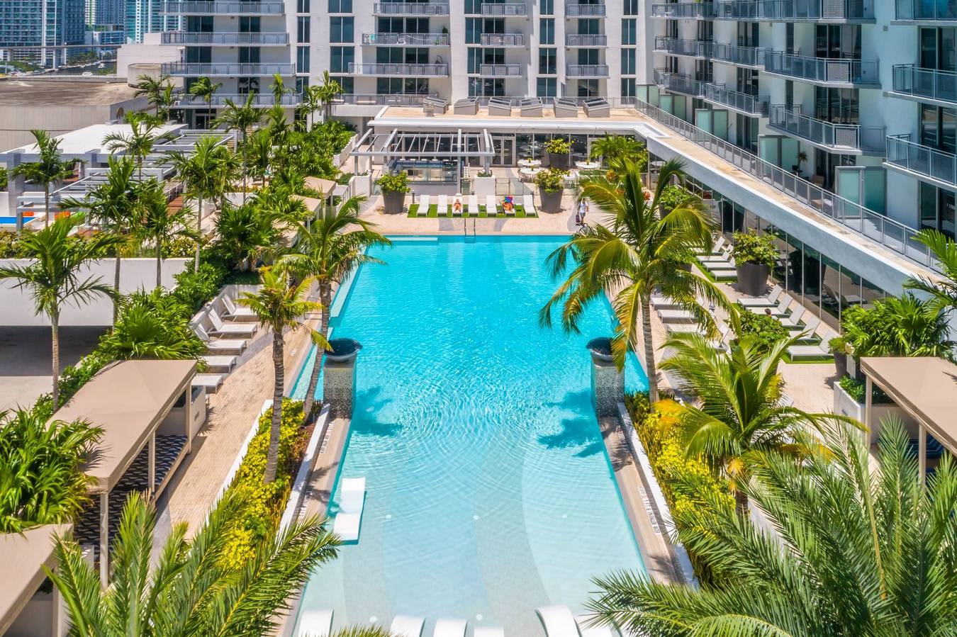 Aerial view of a rectangular swimming pool with turquoise water, flanked by palm trees and lounge chairs, surrounded by contemporary residential towers.