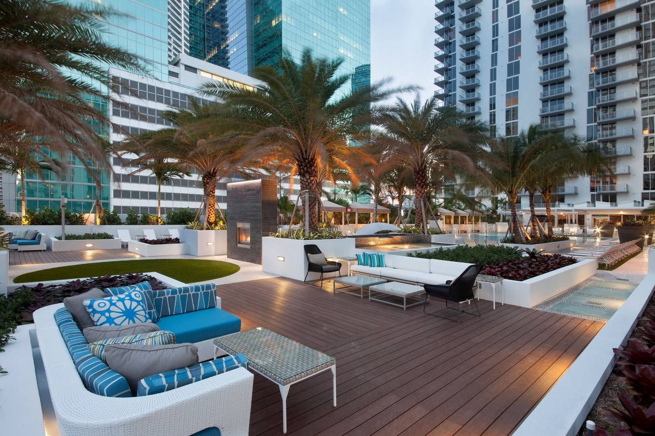 Rooftop terrace with curved white seating, blue cushions, palm trees, and downtown Miami high-rises at dusk.