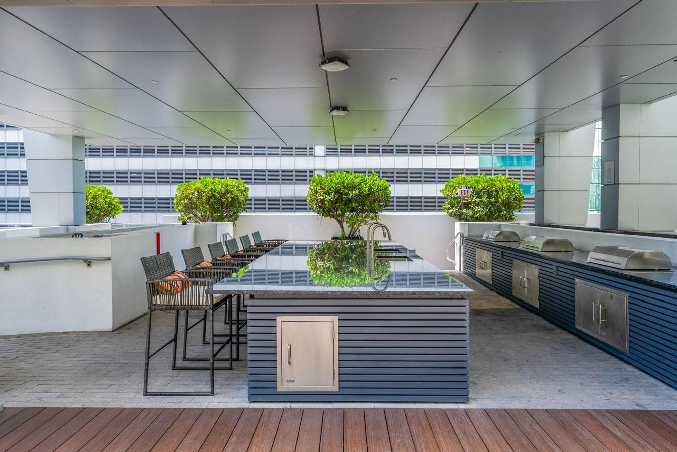 Open-plan kitchen with central island, bar seating, potted topiaries, stainless steel appliances, and city views through large windows.