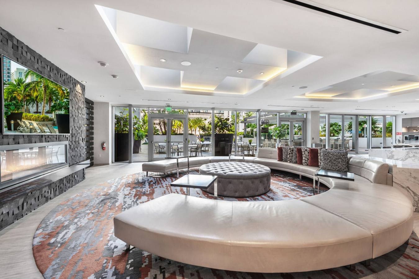 Contemporary lounge with curved cream seating, patterned area rug, fireplace, and floor-to-ceiling windows overlooking tropical outdoor space.