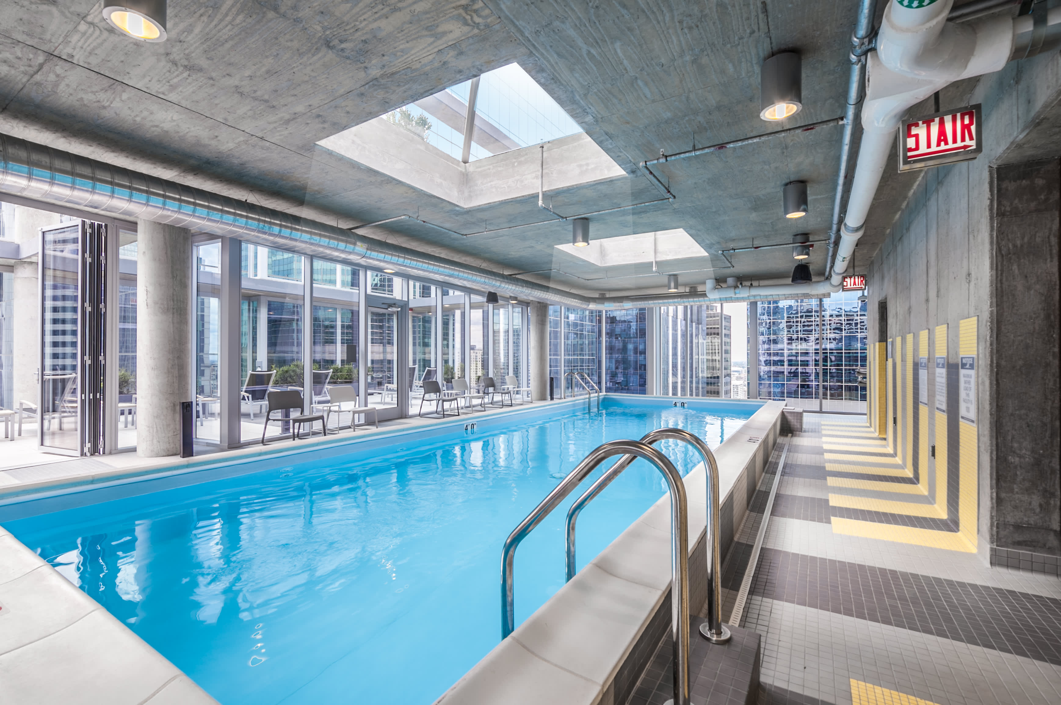 Indoor swimming pool with bright blue water, metal ladder in foreground, floor-to-ceiling windows with city views, skylights, and concrete ceiling with exposed pipes.