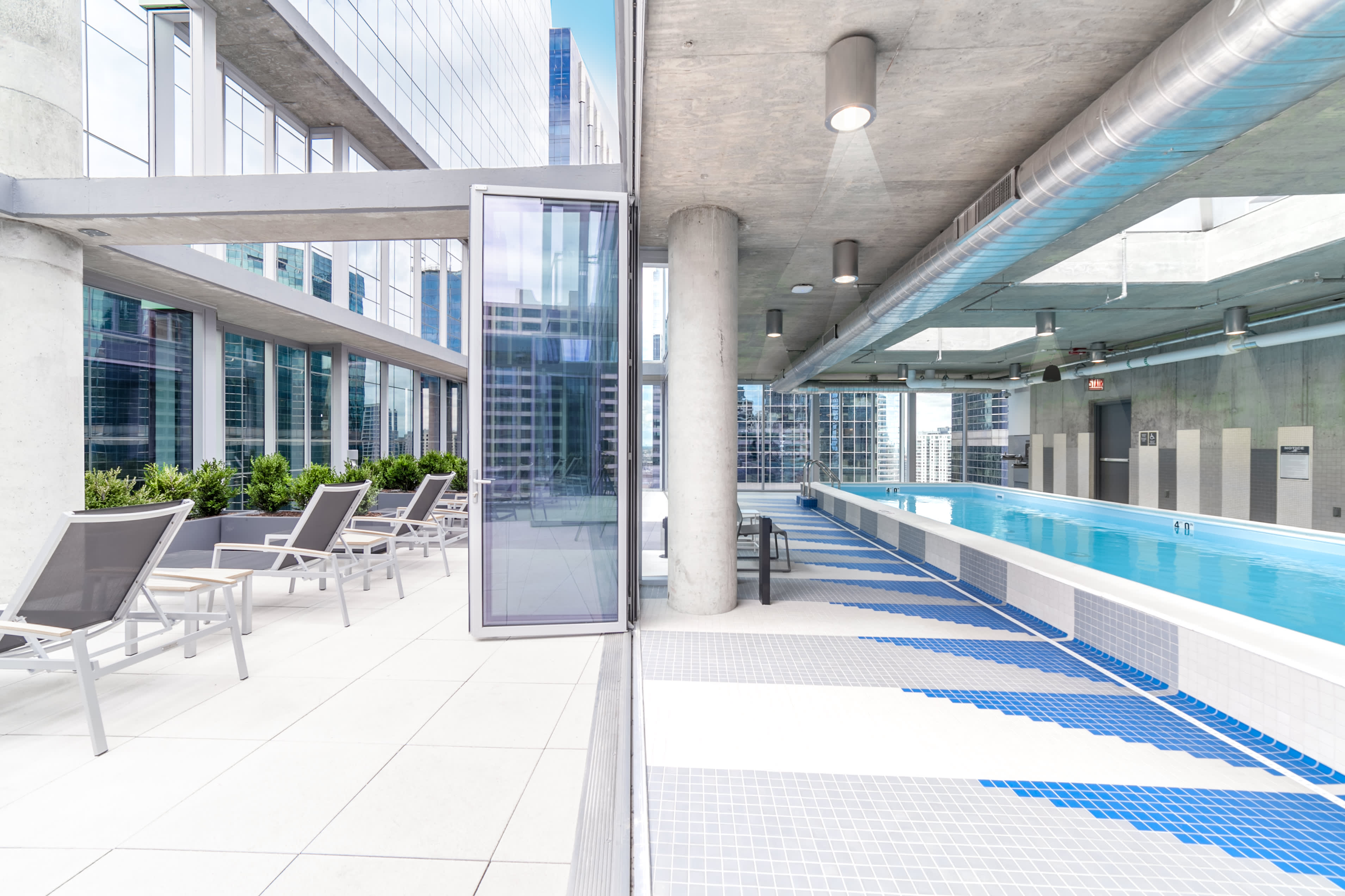 Indoor swimming pool with blue lane markers, concrete columns, and glass doors opening to a terrace with lounge chairs and city views.