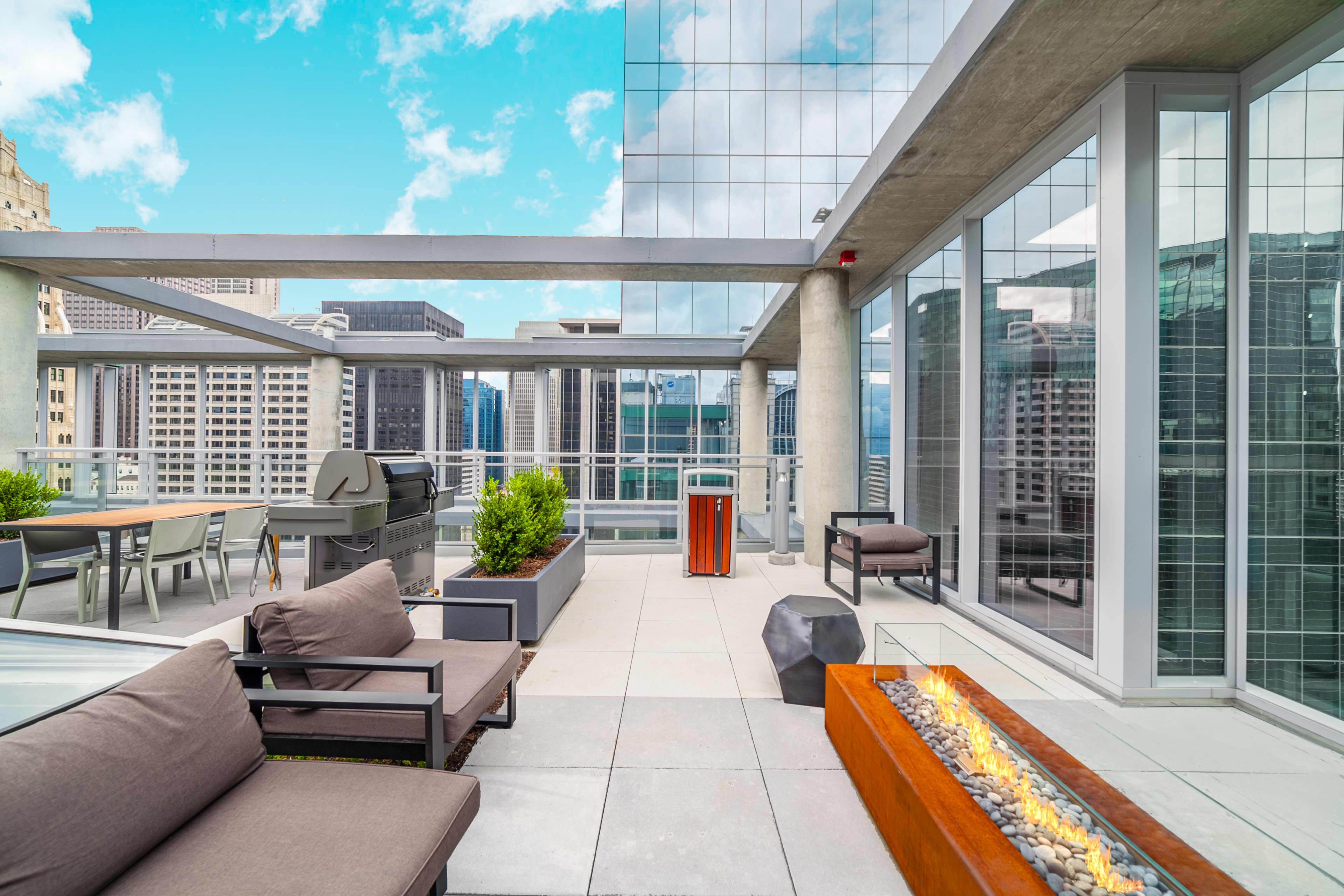 Rooftop terrace with gray lounge seating, linear fire pit, potted plants, grill, and floor-to-ceiling windows with downtown Chicago skyline views.
